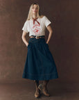 A woman stands against a brown background, wearing The Great Inc.'s The Boxy Crew 100% cotton graphic T-shirt, paired with a red bandana, high-waisted dark denim midi skirt, belt, and brown lace-up boots. Her hands rest in her skirt pockets.