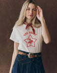 A woman with long blonde hair wears The Boxy Crew t-shirt by The Great Inc., featuring a red star and cowboy graphic, styled with a red bandana, high-waisted blue jeans, and a decorative belt, posing before a brown background.