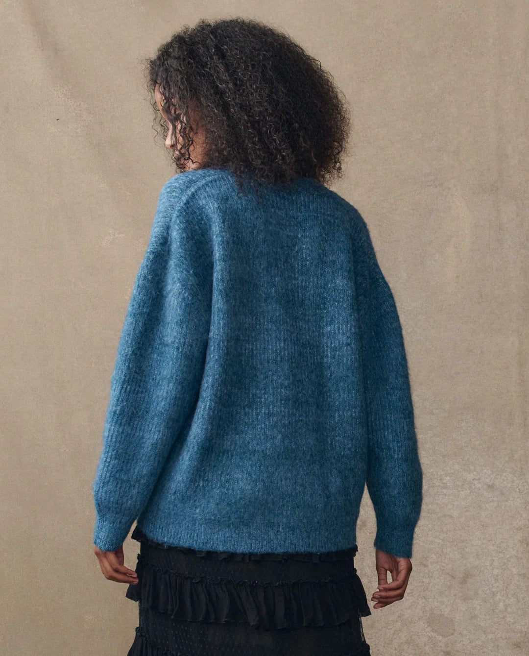 A person with curly hair, seen from behind, wears The Brushed Melange Cardigan by The Great Inc. in blue and a black ruffled skirt, standing against a neutral beige background.