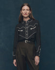 A woman with straight brown hair stands against a dark blue background, smiling softly at the camera while wearing The Western Shirt by The Great Inc.—a black shirt with white piping, arrow details, and pearl snaps—paired with high-waisted dark pants and a belt.