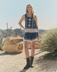 A woman stands in a desert landscape wearing The Sparrow Tank by The Great Inc.—a blue embroidered cotton top with white lace trim—paired with black cowboy boots, surrounded by desert plants and rocks under a clear sky.