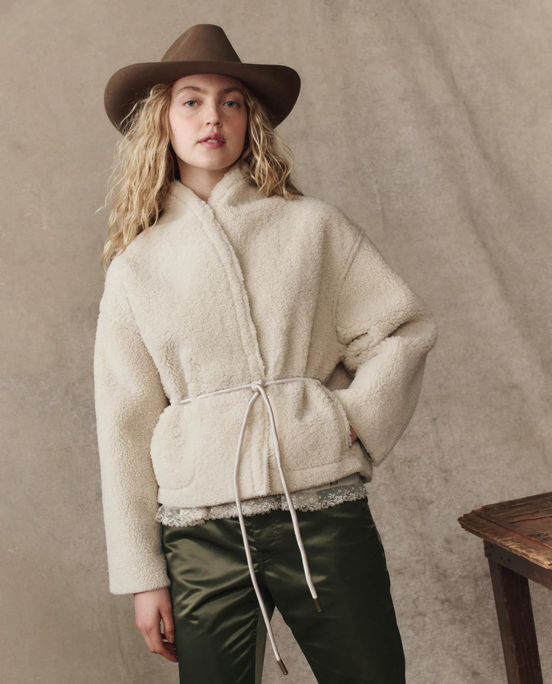 A person with long blonde hair wears The Reversible Drover Coat by The Great Inc.—a cream faux shearling jacket with a drawstring waist—paired with dark green pants and a brown wide-brimmed hat, standing before a neutral textured background.