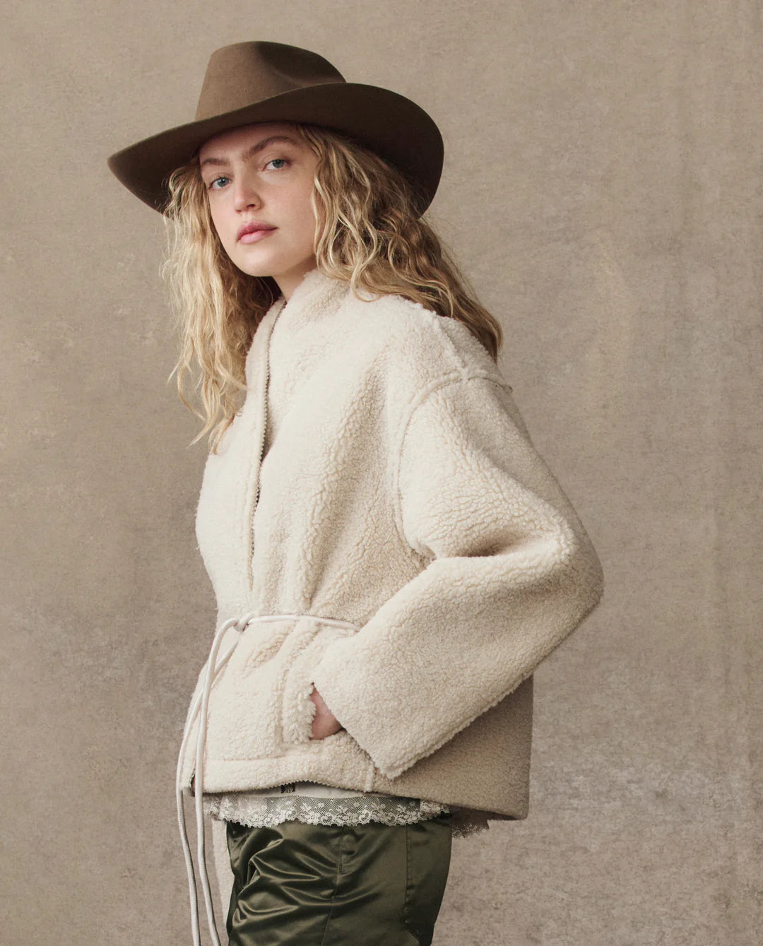 A person with long blonde hair models The Great Inc.’s Reversible Drover Coat, cream-colored and tied at the waist, paired with olive green pants and a brown wide-brimmed hat, standing before a beige textured background.
