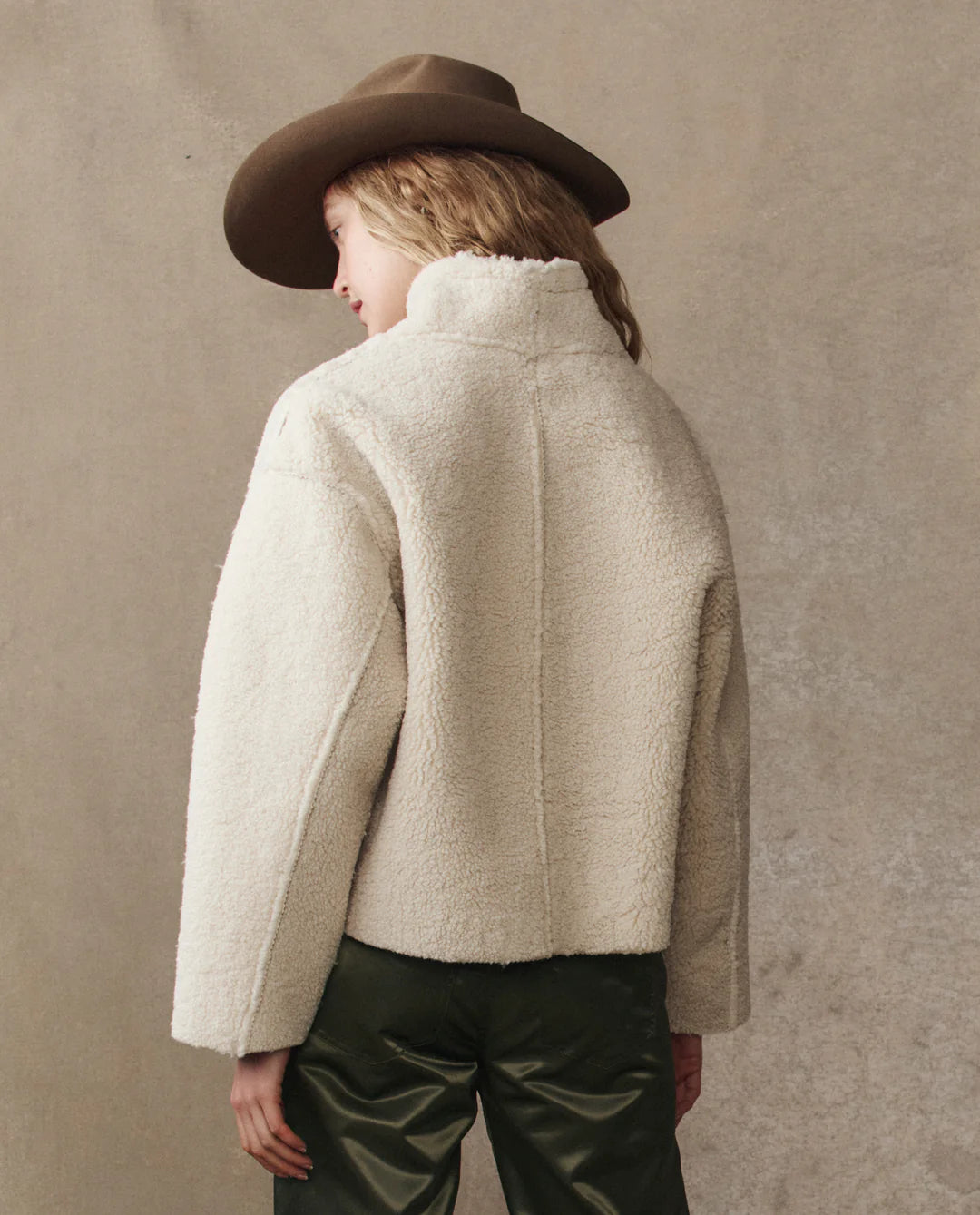A person wearing The Great Inc.'s Reversible Drover Coat in off-white faux shearling and a brown wide-brimmed hat with dark green pants stands facing away from the camera against a neutral, textured background.