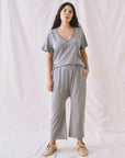 A woman poses in The Great Inc.'s The Jersey Crop outfit, featuring a gray V-neck T-shirt paired with elastic waist jersey pants that have a wide straight leg and dropped rise. She stands against a plain, light backdrop, wearing casual slip-on shoes.