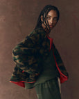 A person with braided hair poses before a brown backdrop, wearing The Great Inc. Reversible Plush Bomber with vintage camo fleece, red lining, and green clothing underneath.