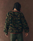 A person with braided hair faces away, wearing The Great Inc. Reversible Plush Bomber in camo fleece against a textured brown background.