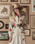 A woman wearing a beige embroidered outfit with red flowers and The Great Inc. Leather Concho Belt stands before a wall of framed horse paintings, sporting a tan cowboy hat and looking at the camera.