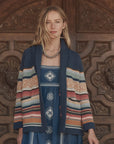 A woman with long blonde hair stands before an ornate wooden door in a blue patterned dress and The Great Inc.'s Mountain Lodge Cardigan, a chunky knit with navy, orange, cream, and red stripes. She accessorizes with a beaded necklace and serene expression.