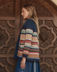A woman in profile stands before ornate wooden doors, wearing The Mountain Lodge Cardigan by The Great Inc., a Southwestern-inspired piece with blue, orange, and cream horizontal stripes over a blue dress.
