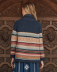 A woman with light brown hair faces a carved wooden door, wearing The Mountain Lodge Cardigan by The Great Inc., which features colorful horizontal stripes, paired with a patterned blue skirt.