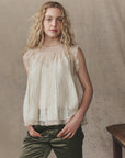 A young woman with wavy blonde hair wears The Keepsake Lace Shell Top by The Great Inc. with olive green pants, standing against a neutral, textured background and facing the camera with a relaxed expression.