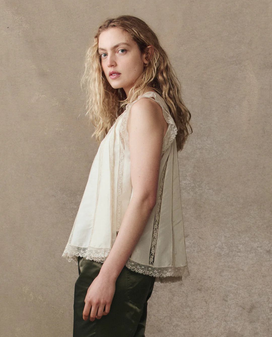 A woman with wavy blonde hair stands sideways against a beige background, wearing The Keepsake Lace Shell Top by The Great Inc. in off-white, paired with dark green pants.