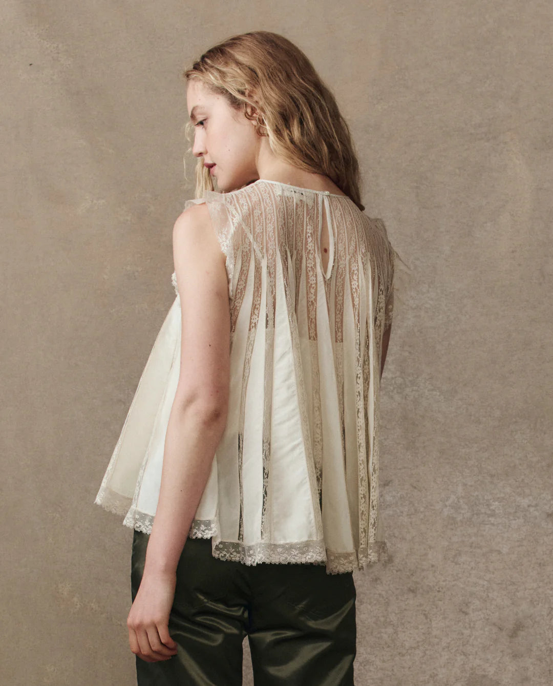 A woman with wavy blonde hair stands sideways, wearing The Keepsake Lace Shell Top by The Great Inc.—a sleeveless lace top with pleated panels, a keyhole back, and romantic Victorian-inspired details—paired with dark green pants.