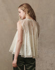 A woman with wavy blonde hair stands sideways, wearing The Keepsake Lace Shell Top by The Great Inc.—a sleeveless lace top with pleated panels, a keyhole back, and romantic Victorian-inspired details—paired with dark green pants.