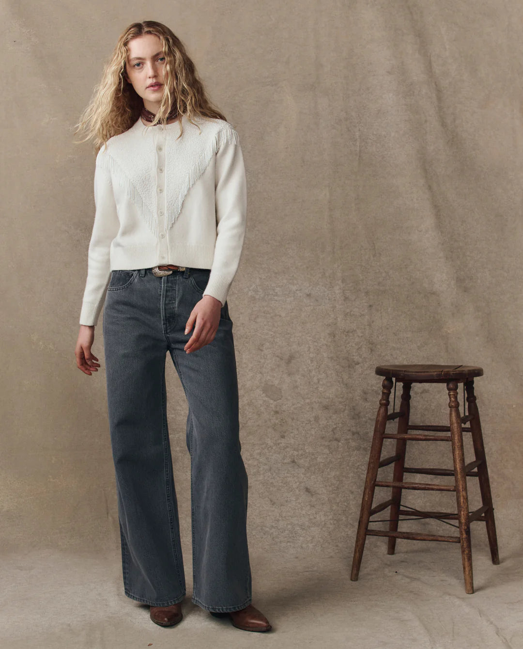 A person with long wavy blonde hair wears The Beaded Fringe Cardigan by The Great Inc., paired with dark high-waisted wide-leg jeans and brown shoes, standing beside a wooden stool against a neutral beige backdrop.