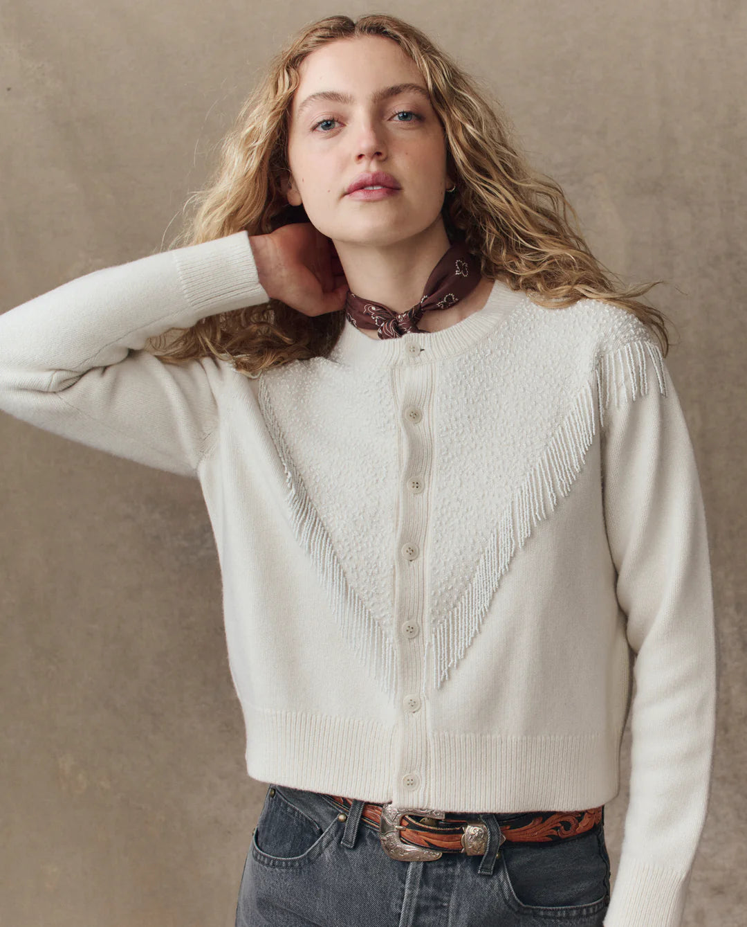 A young woman with long curly blonde hair wears The Beaded Fringe Cardigan by The Great Inc. in a cream wool-cotton blend, styled with dark jeans, a brown belt, and a brown neck scarf, standing against a beige background.