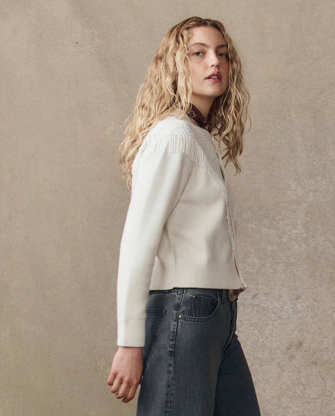 A young woman with long, wavy blonde hair stands sideways against a plain beige background, wearing The Great Inc.'s Beaded Fringe Cardigan in white and dark high-waisted jeans.