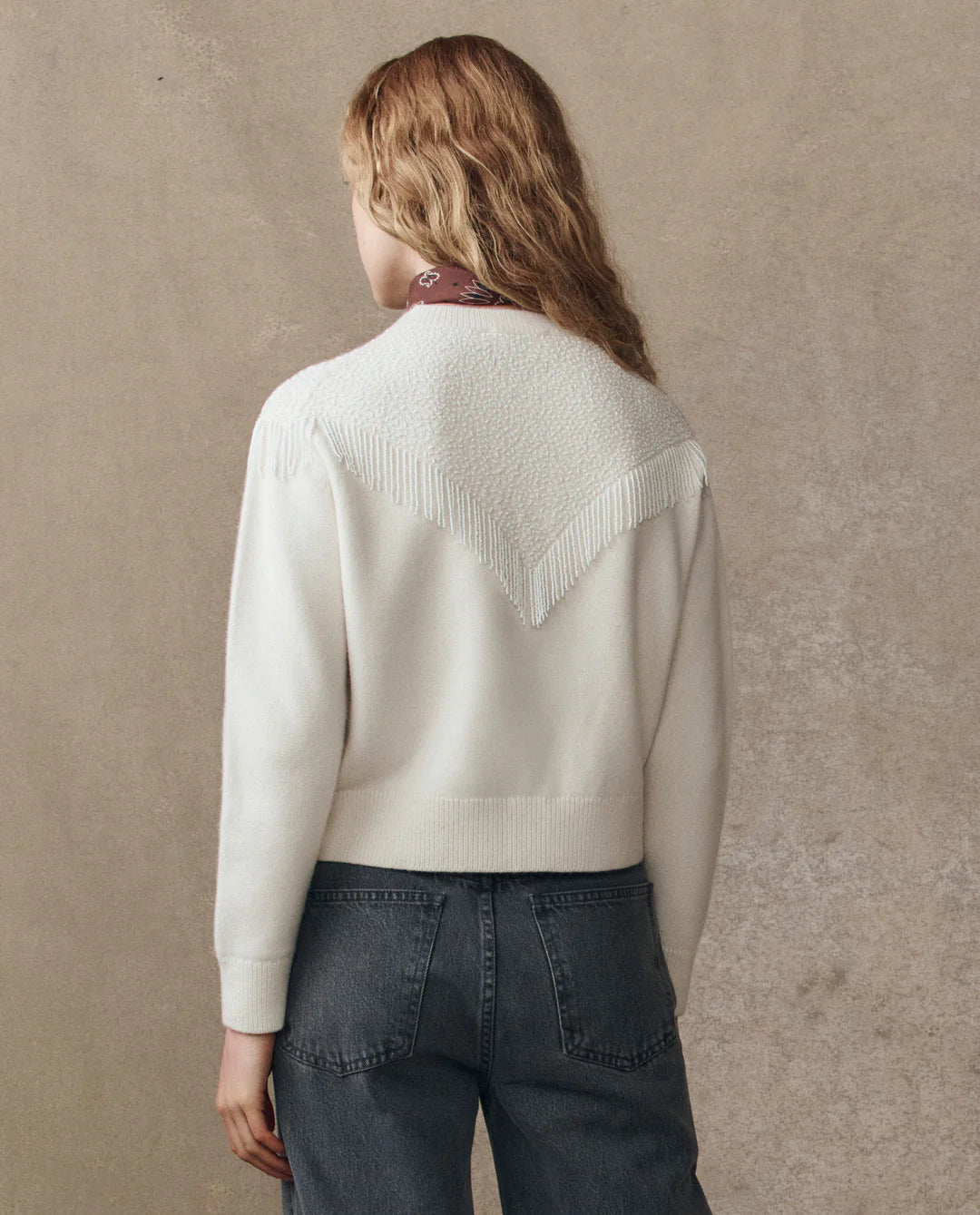 A person with wavy, light brown hair is seen from behind wearing The Beaded Fringe Cardigan by The Great Inc. with textured detailing and blue jeans, standing against a beige textured wall.