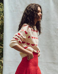 A woman with long, dark hair stands sideways, wearing The Heart Crew by The Great Inc., paired with a red skirt. She is outdoors against a plain backdrop, with greenery visible at the side.