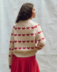 A person with curly hair is facing away, wearing The Heart Crew by The Great Inc., a pullover crewneck sweater with red heart patterns, paired with a red skirt. The background is a plain light-colored wall.