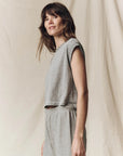 A woman with wavy brown hair wears The Square Tee by The Great Inc., a light gray sleeveless crop top, paired with matching high-waisted denim, standing sideways and smiling softly against a neutral beige fabric background.