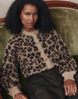 A woman with curly hair sits on a green velvet sofa, wearing The Windsor Cardigan by The Great Inc.—a beige and black leopard jacquard cropped knit—paired with dark pinstriped pants, gazing relaxedly at the camera.