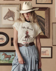 A woman in a cowboy hat, graphic tee, denim skirt, and The Great Inc. Tooled Waist Belt stands before a wall of horse art, her head tilted so the hat shades part of her face.