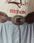 A close-up of a person in a beige shirt with "STETSON" in red, wearing The Great Inc.'s Tooled Waist Belt with an ornate silver buckle, paired with a light blue buttoned skirt.