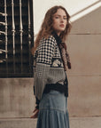 The Pony Cardigan by The Great Inc. styled with a long denim skirt and scarf beside a beige wall and window.