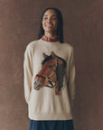 A woman with braided hair smiles in The Great Inc. Stable Pullover, an oversized crew sweater with a large horse intarsia graphic. She pairs it with a red patterned neck scarf, standing against a brown background.