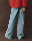 A person wearing The Great Inc. Slouch Wide Leg jean in light blue and a red jacket stands with their back turned against a dark brown background, left arm relaxed at their side.