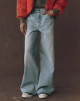 A person wearing The Great Inc. Slouch Wide Leg jean in light blue, a green top, red jacket, and white sneakers stands against a plain brown background with hands in pockets and face not visible.