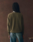 A person with braids wears The College Sweatshirt by The Great Inc., a green vintage style featuring a Wild Panther graphic.