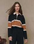 A woman with long brown hair wears The Rugby Sweater by The Great Inc.—a boxy knit black pullover with wide brown and white stripes—standing against a plain beige background, looking at the camera and smiling slightly.