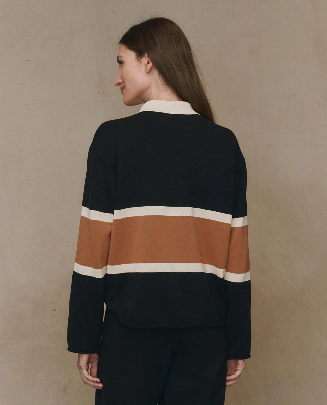 A person with long brown hair stands with their back to the camera, wearing The Rugby Sweater by The Great Inc.—a boxy knit top featuring wide horizontal brown and white stripes—set against a plain beige background.