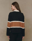 A person with long brown hair stands with their back to the camera, wearing The Rugby Sweater by The Great Inc.—a boxy knit top featuring wide horizontal brown and white stripes—set against a plain beige background.