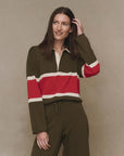 A woman with long brown hair smiles and touches her hair while wearing The Rugby Sweater by The Great Inc.—an olive green boxy top with a red and white horizontal chest stripe—posing against a neutral background.