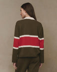 A woman with long brown hair stands with her back to the camera, wearing The Great Inc.'s The Rugby Sweater in olive green with a wide red and white stripe across the back, paired with matching pants against a beige background.