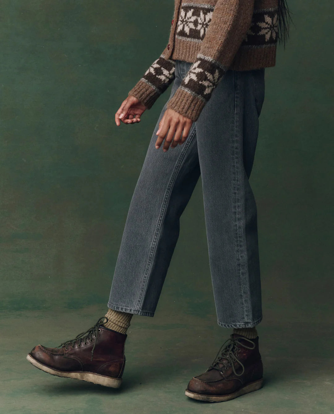 A person wears The Wayne Jean by The Great Inc., paired with a brown patterned sweater, olive socks, and dark brown lace-up boots. Only their lower half is visible against a green background.