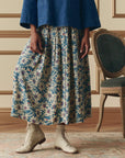A person stands indoors wearing The Highland Skirt by The Great Inc.—a long floral pleated skirt with yellow and white flowers and an elastic waistband—paired with a blue top and white lace-up ankle boots beside a vintage green chair.