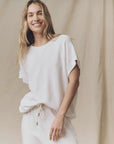 A person with long blonde hair smiles in front of a beige fabric backdrop, wearing The Wedge Sweatshirt by The Great Inc., effortlessly channeling a relaxed, vintage-inspired vibe.
