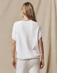 A woman with long, wavy blonde hair stands with her back to the camera, wearing The Wedge Sweatshirt by The Great Inc. and matching white pants against a beige fabric backdrop.