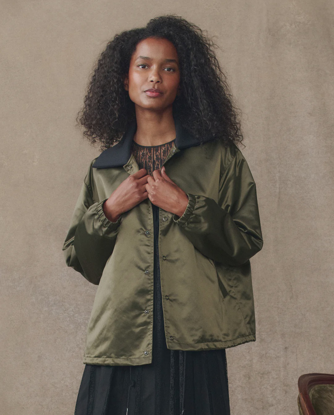 A woman with long curly hair wears The Great Inc.'s The Satin Coach's Jacket in olive green, featuring an oversized rib collar, layered over a dark blouse against a neutral beige background.