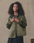 A woman with long curly hair wears The Great Inc.'s The Satin Coach's Jacket in olive green, featuring an oversized rib collar, layered over a dark blouse against a neutral beige background.