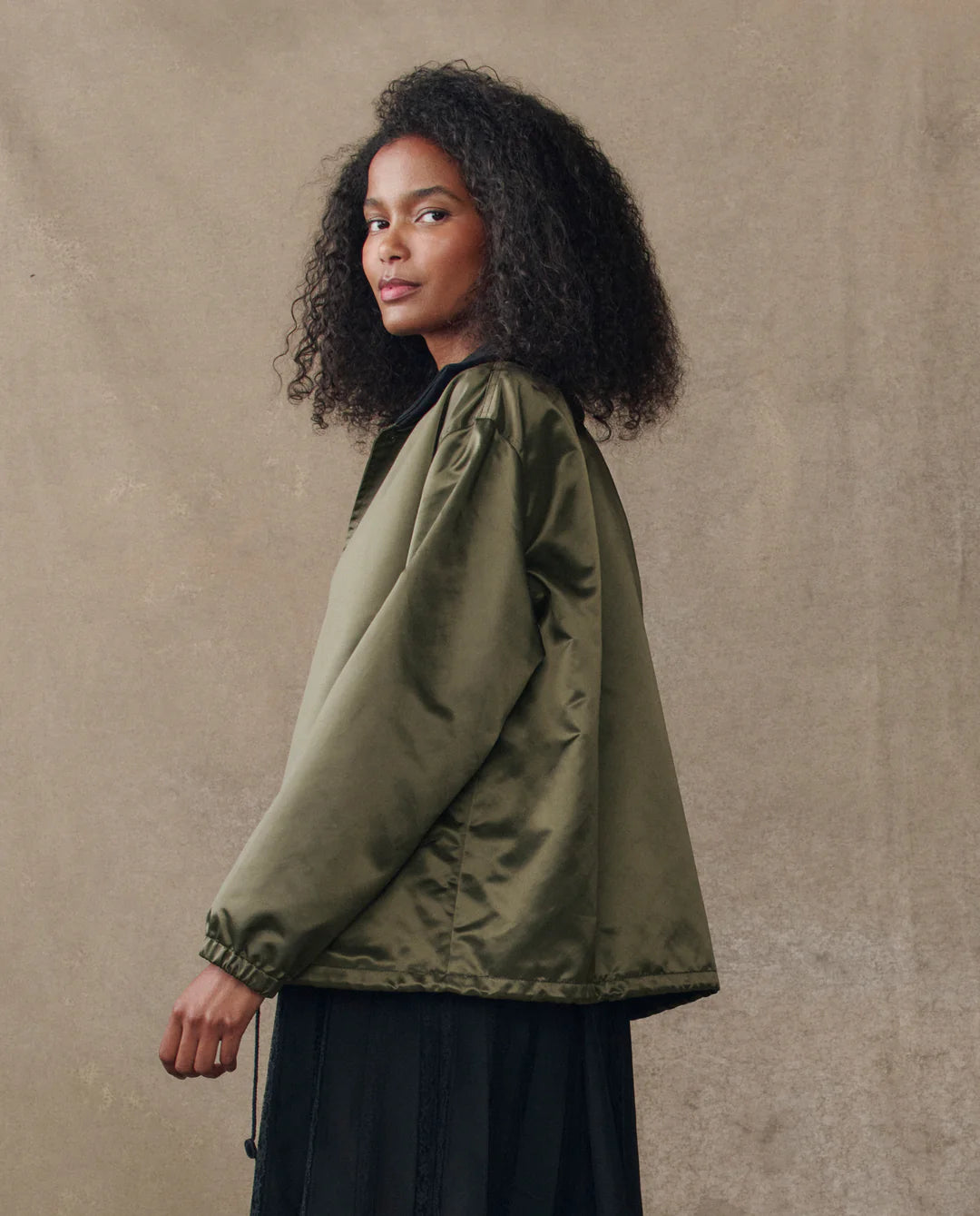 A person with curly hair stands in profile against a neutral background, wearing The Satin Coach's Jacket in olive green by The Great Inc. and a black skirt.