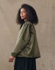 A person with curly hair stands in profile against a neutral background, wearing The Satin Coach's Jacket in olive green by The Great Inc. and a black skirt.