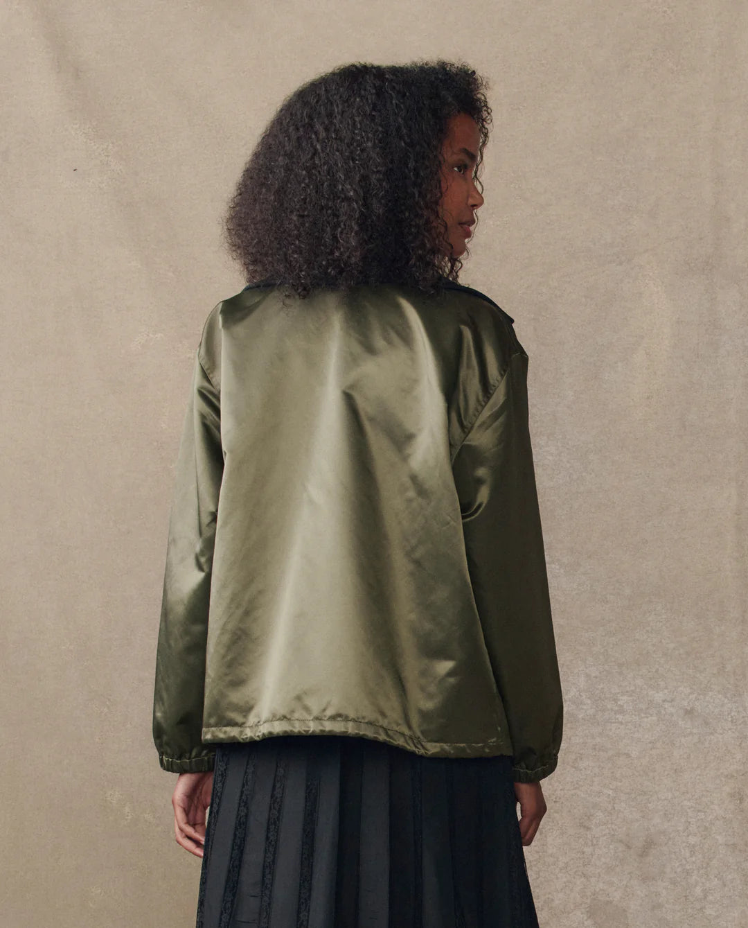 A woman with curly hair faces away, wearing The Great Inc.'s Satin Coach's Jacket in shiny olive green with an oversized rib collar and a black skirt, set against a neutral beige background.