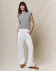 A woman stands against a beige backdrop in The Great Inc.'s black-and-white Peak Shoulder Tee with a crewneck fit, white wide-leg pants, and tan moccasins—her effortlessly relaxed look channels handcrafted Los Angeles style.