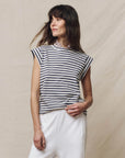 A woman with long brown hair wears The Peak Shoulder Tee by The Great Inc.—a handcrafted navy and white crewneck—paired with white pants, standing against a neutral fabric backdrop while looking to the side and holding her shirt’s hem.